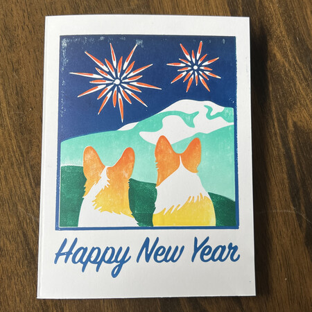 A card with the words Happy New Year below hand printed artwork of two corgis looking at fireworks across a field with mountains in the background